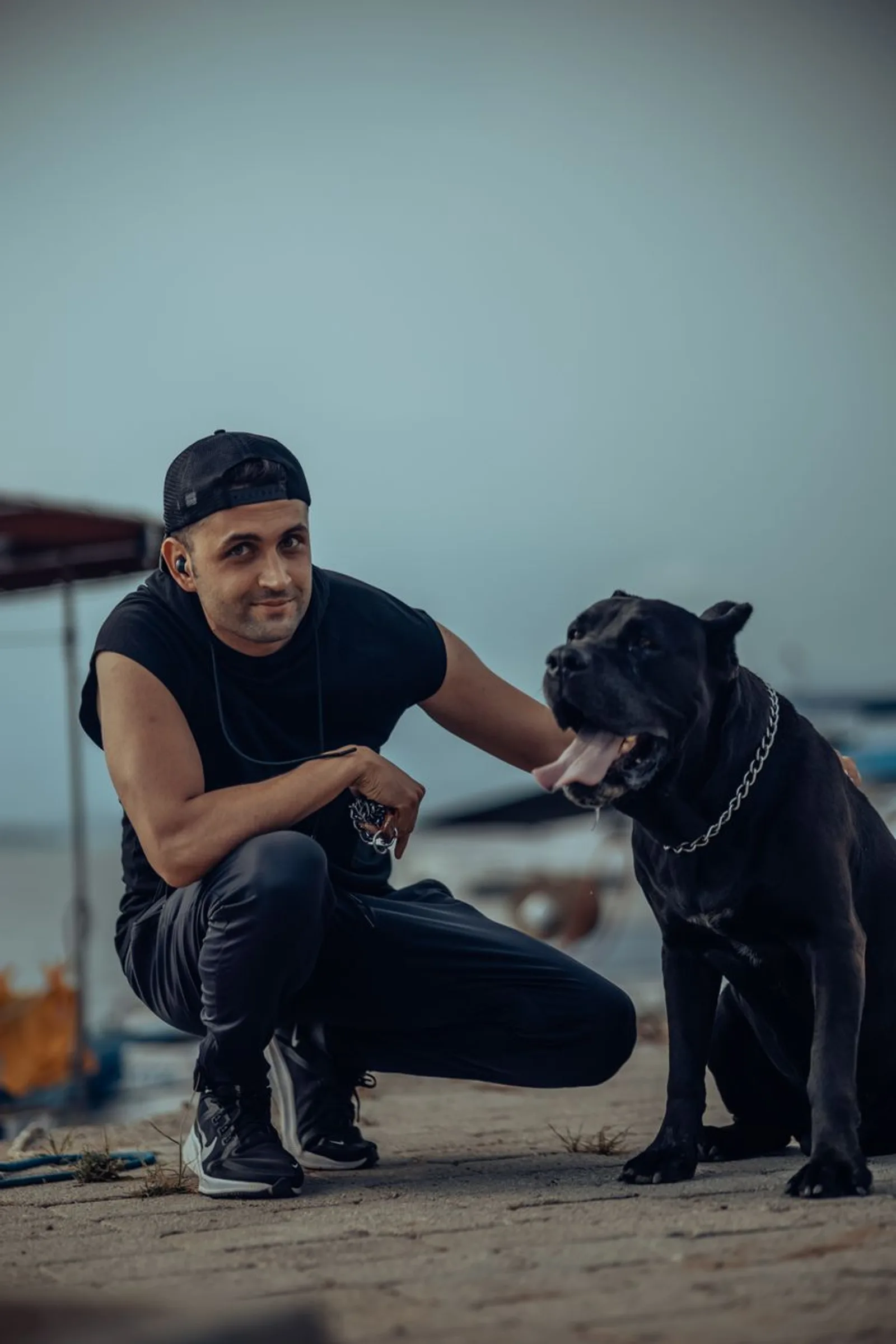 Homem agachado ao lado de um Cane Corso preto com guia, em uma praia